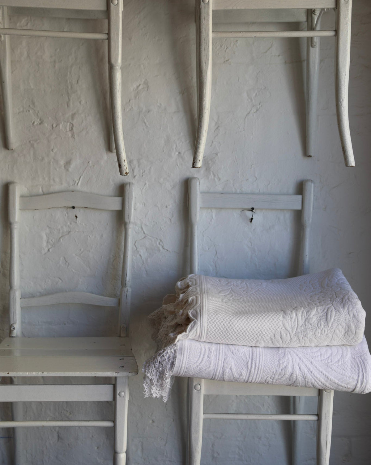 A stack of white upholstered chairs arranged against a textured white wall, with a folded white lace blanket or throw resting on the bottom-right chair.