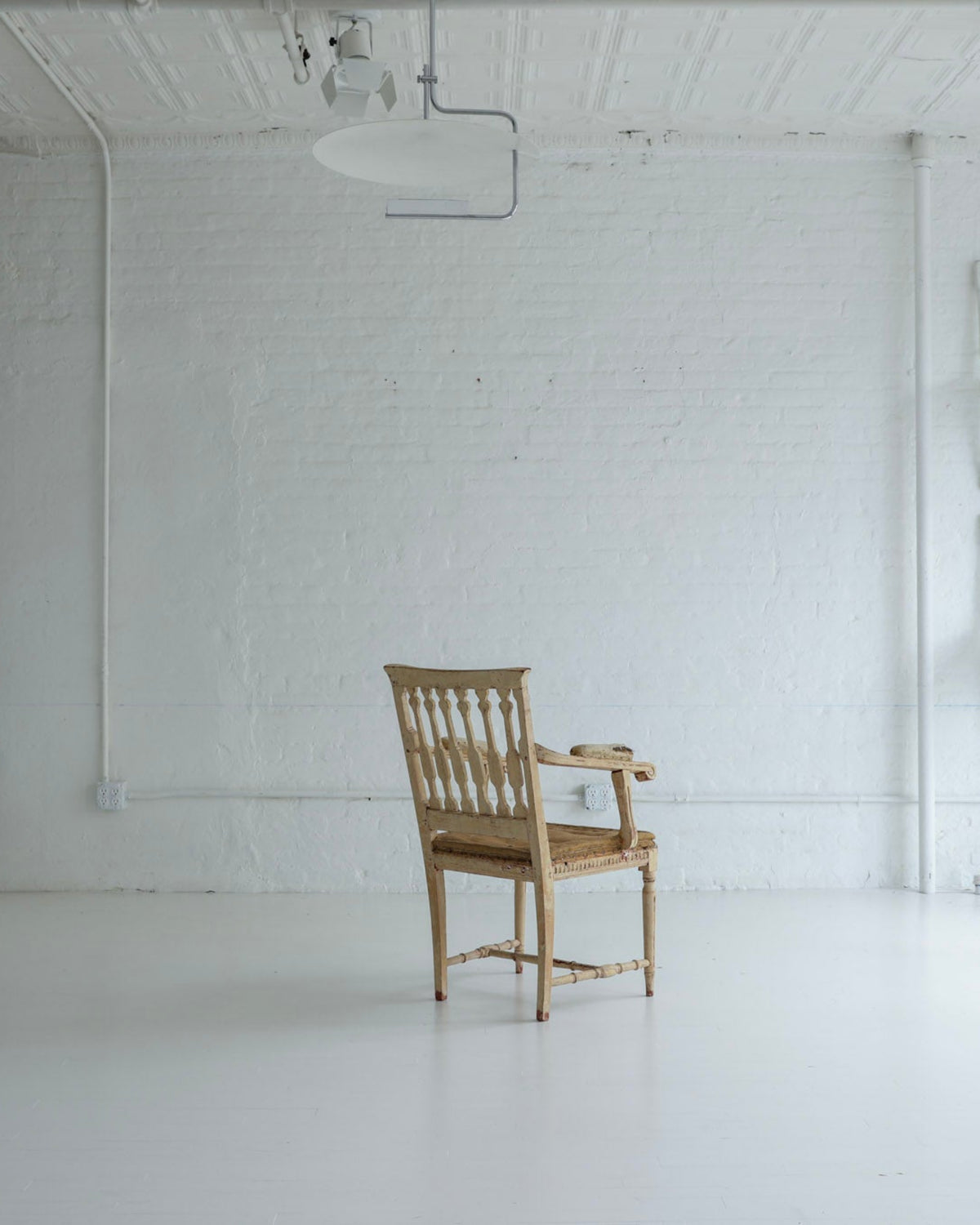 A single wooden armchair sits centered in a bright, white studio or gallery space with white brick walls, track lighting, and additional white chairs mounted on the wall.