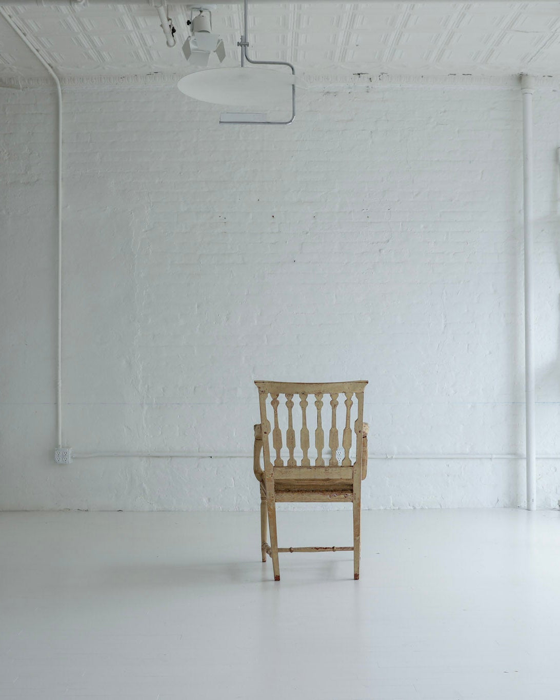A single light wood spindle-back chair stands centered in a bright, white studio or gallery space with a white brick wall, white floor, and several white chairs mounted on the wall to the right.