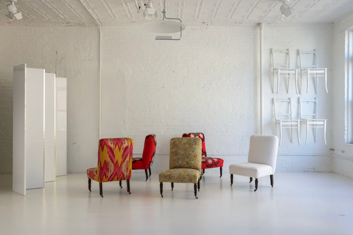 A styled interior room with white walls and floor, featuring several upholstered armchairs and a white chair stacked against the wall. The chairs include bold red, yellow, and floral patterned fabrics, alongside a solid white chair.