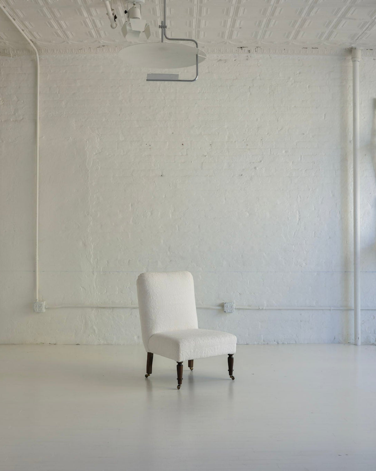 A single upholstered armchair in off-white stands centered in a bright, white studio or gallery space with a textured wall, exposed ceiling beams, and additional white furniture pieces mounted or placed along the left and right walls.