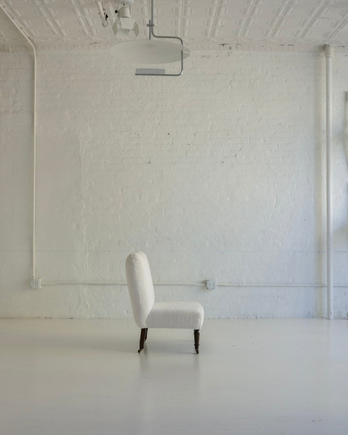 A minimalist white upholstered armchair with dark legs stands in the center of a bright, white-walled studio or gallery space. Several matching white chairs are stacked against the right wall, and a tall white cabinet or screen is visible on the left.