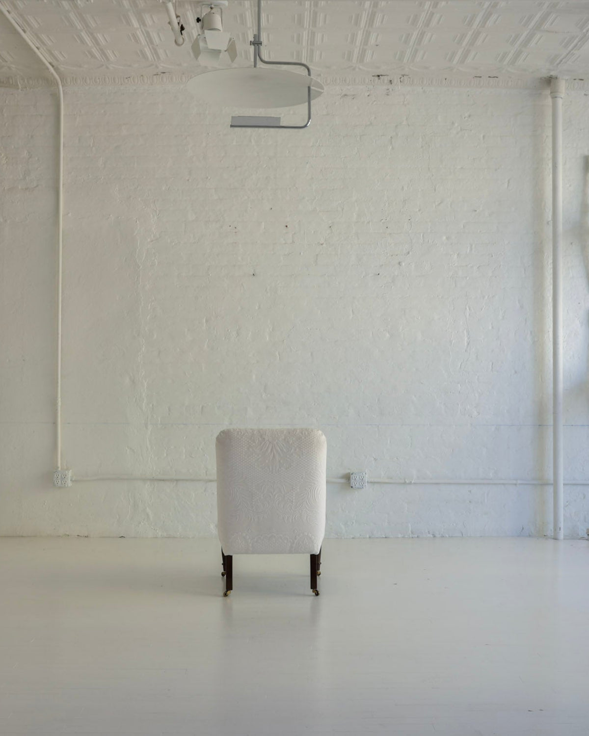 A single upholstered armchair sits centered in a bright, white studio or gallery space with a high ceiling, track lighting, and several white chairs stacked against the right wall.