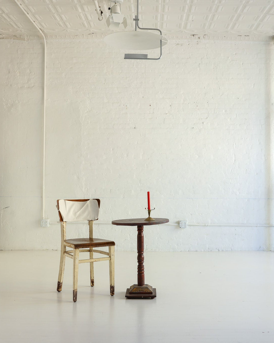 A minimalist interior scene in a bright white room with a small round wooden table and a light wood chair centered near the wall, with additional white chairs mounted on the wall and a tall white cabinet on the left.