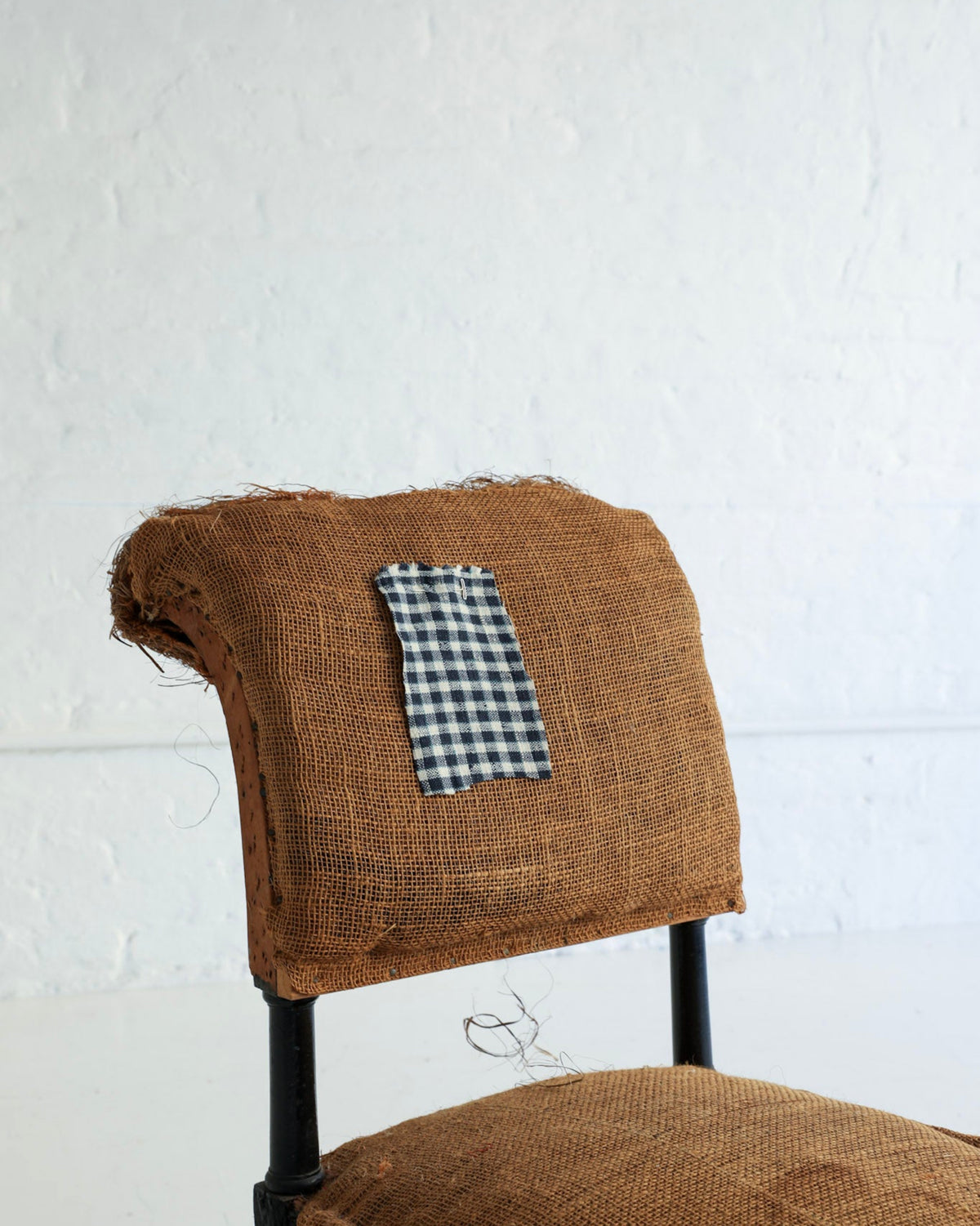 A rustic upholstered chair with a brown burlap-covered backrest and seat cushion, featuring a small black-and-white gingham fabric patch on the backrest, set against a white textured wall.