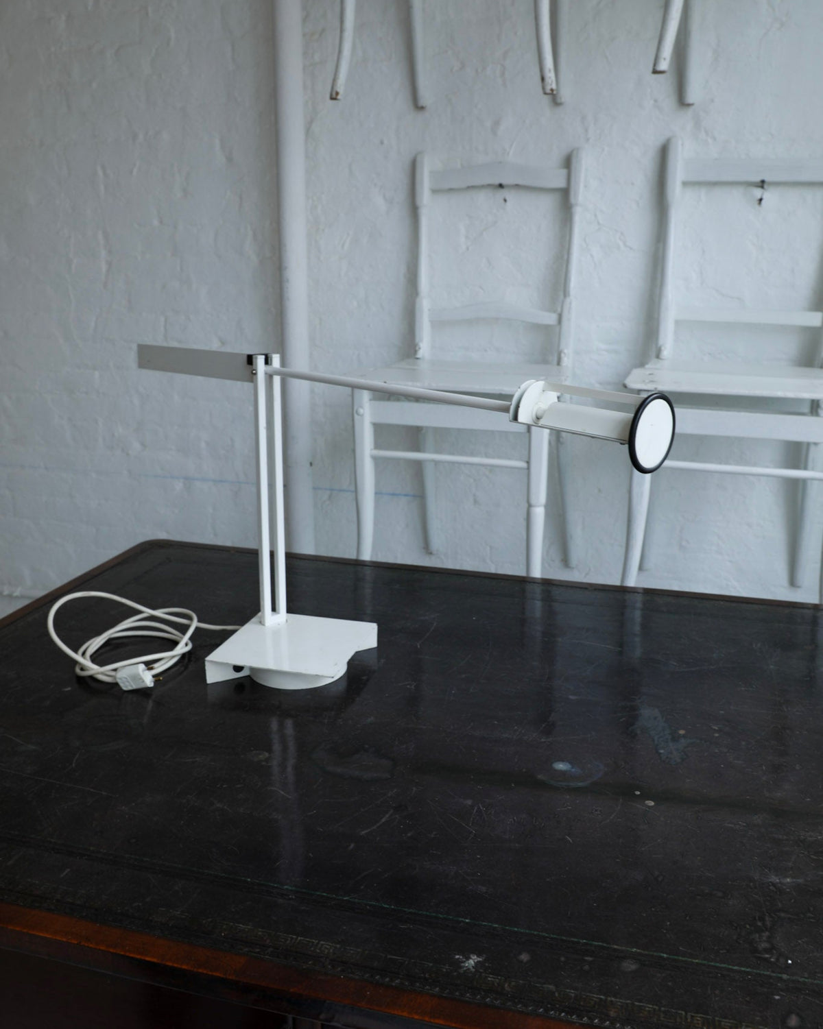 A modern white adjustable desk lamp with a square base and a long horizontal arm, resting on a dark wooden table in a room with white chairs against a white brick wall.