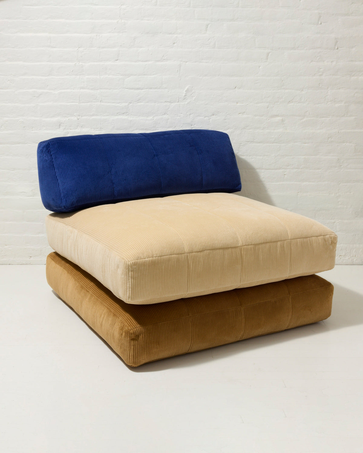 Three cushioned floor chairs in blue, beige, and brown against a white brick wall.