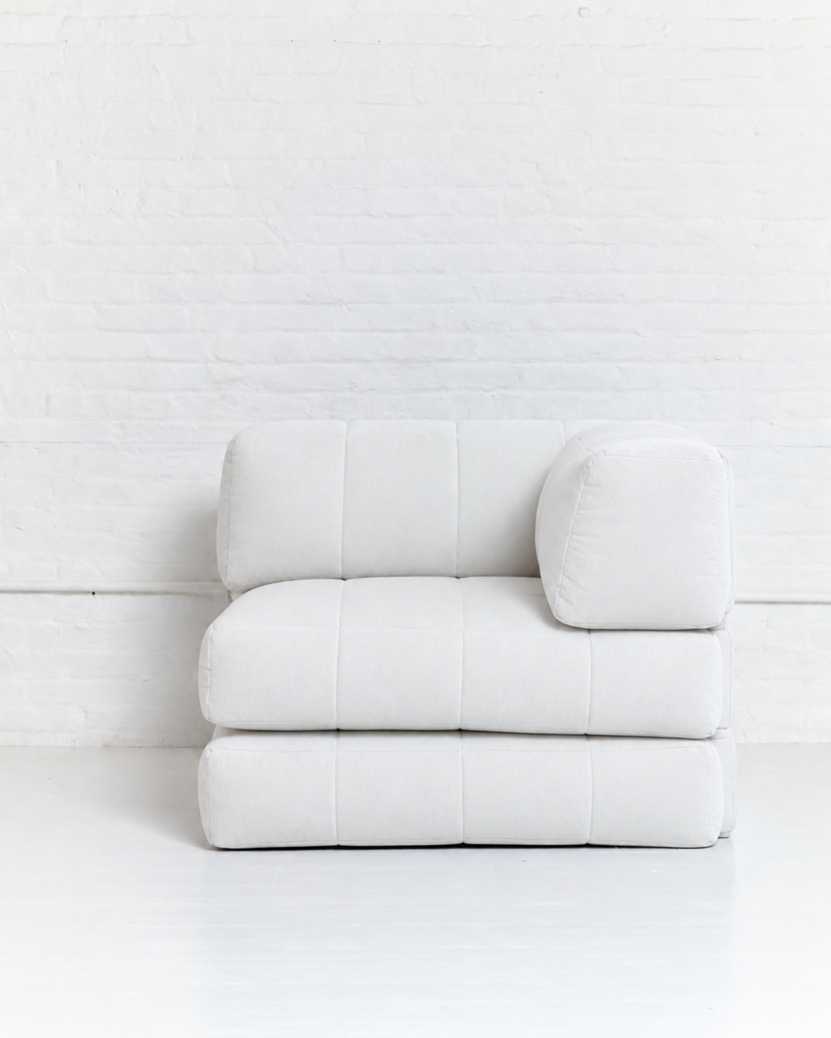 A modern modular white upholstered ottoman or low seat with a square backrest cushion and a separate cylindrical lumbar pillow, set against a white brick wall and white floor.