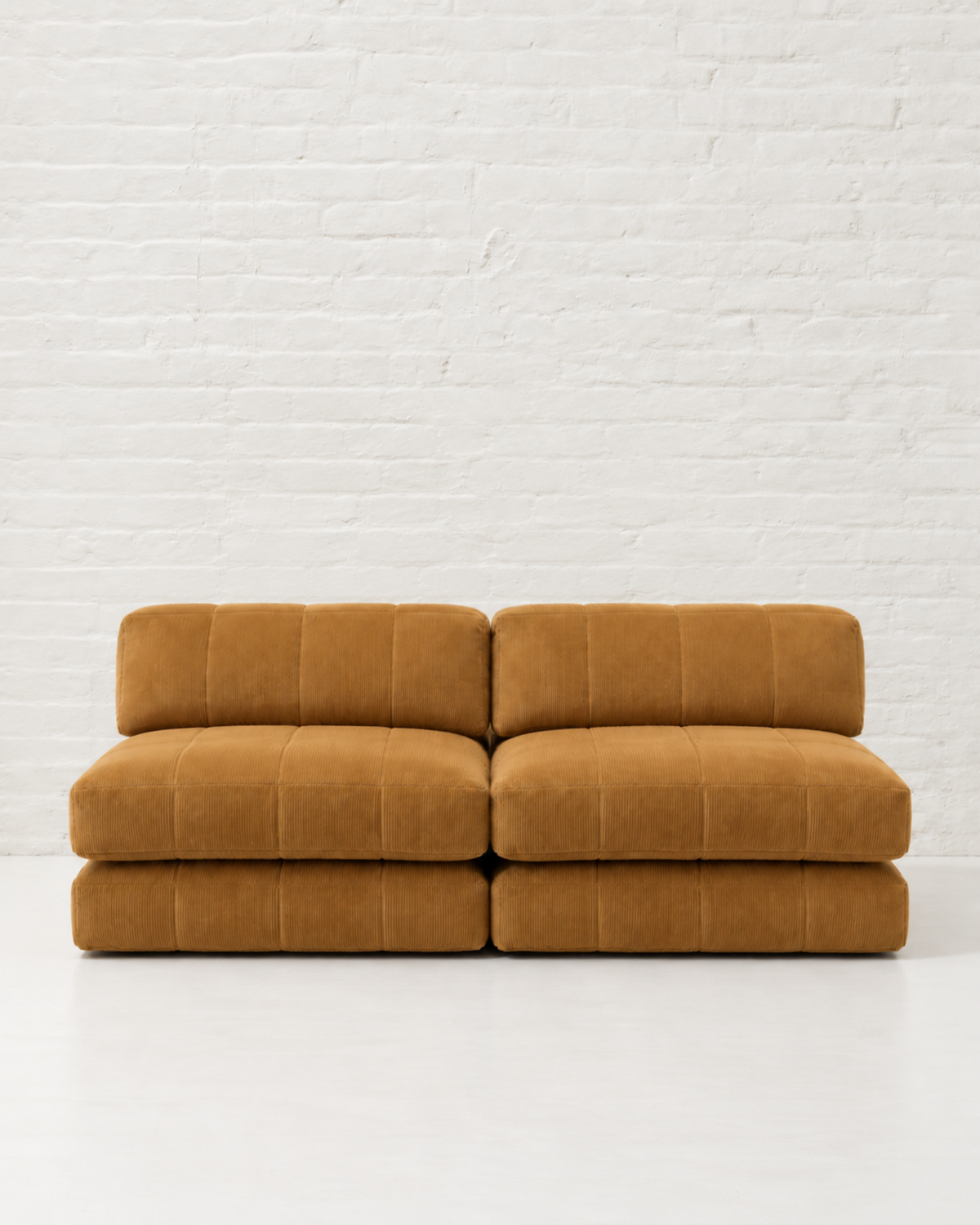 A modern two-seater modular sofa upholstered in a textured mustard-yellow velvet fabric with channel tufting, set against a white brick wall background.