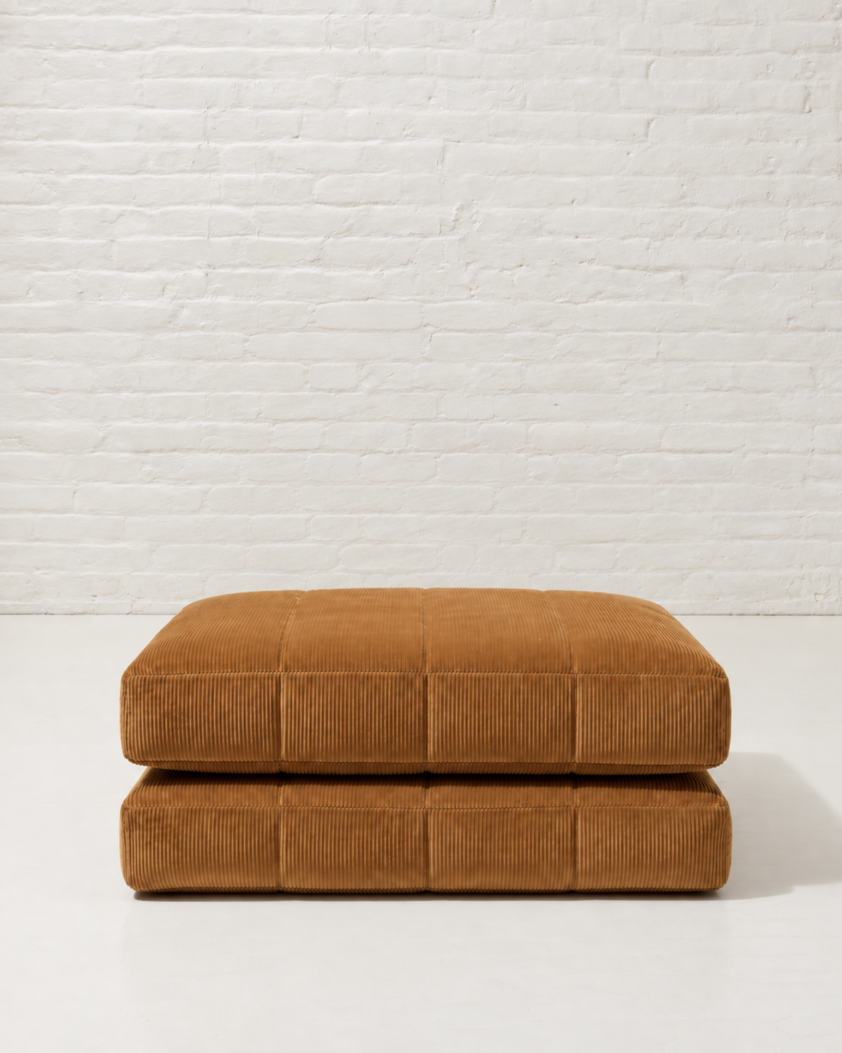 Two stacked square floor cushions in a warm camel corduroy fabric, set against a white brick wall background.