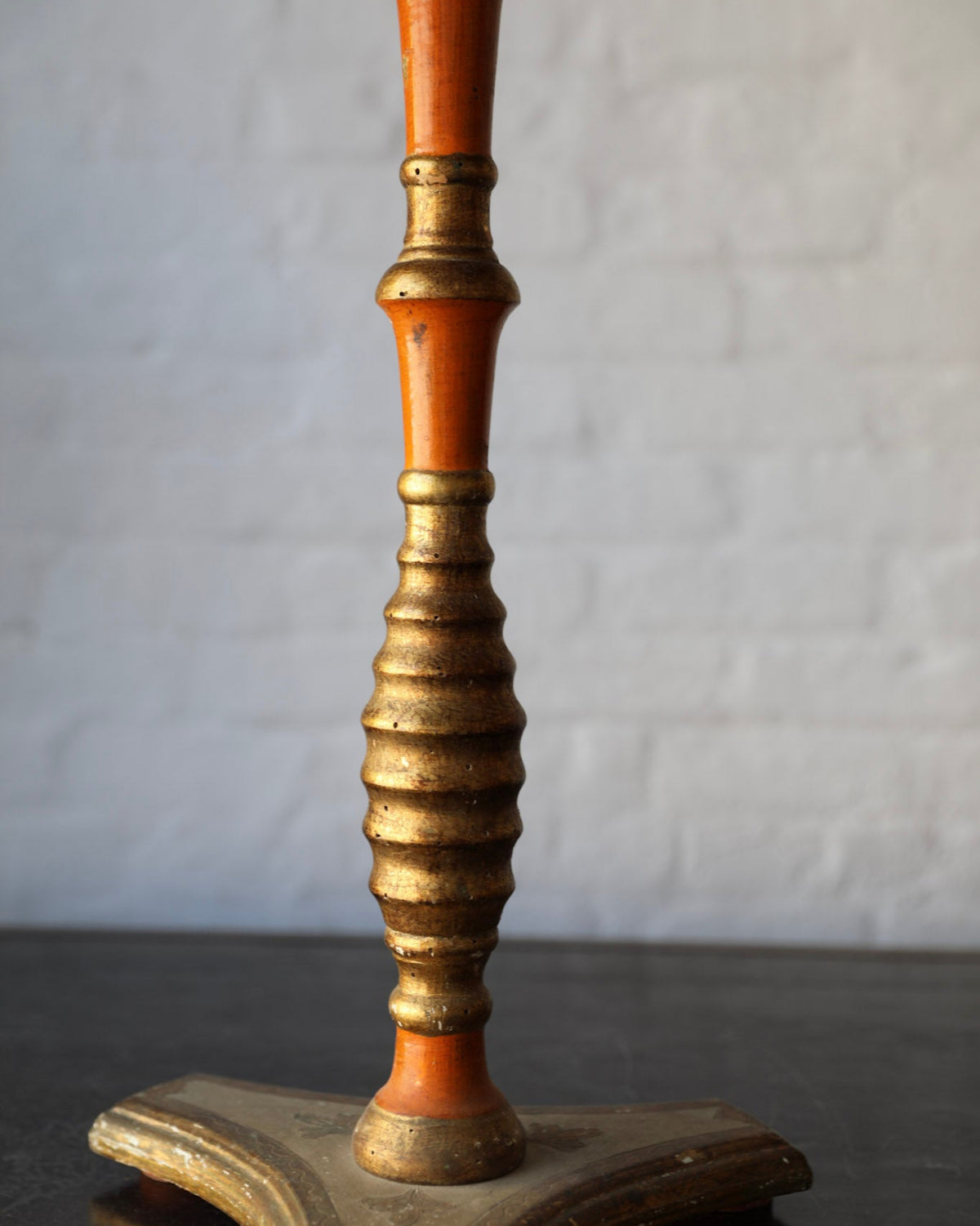 Close-up of a vintage table pedestal featuring a turned orange-brown wood stem with ornate gold gilding and a matching gold-toned base, set against a white brick wall background.