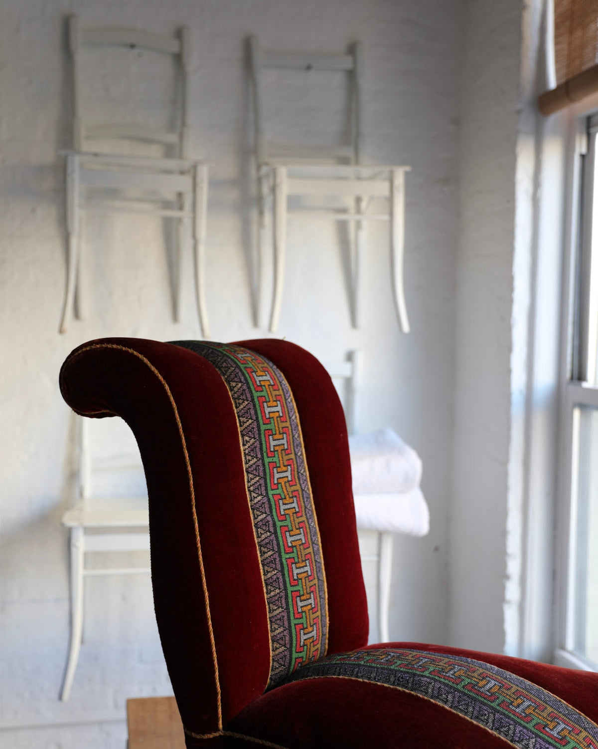 A close-up of a red velvet wingback armchair with a colorful patterned textile panel on the backrest and seat, set against a white brick wall with two white chairs hanging in the background.