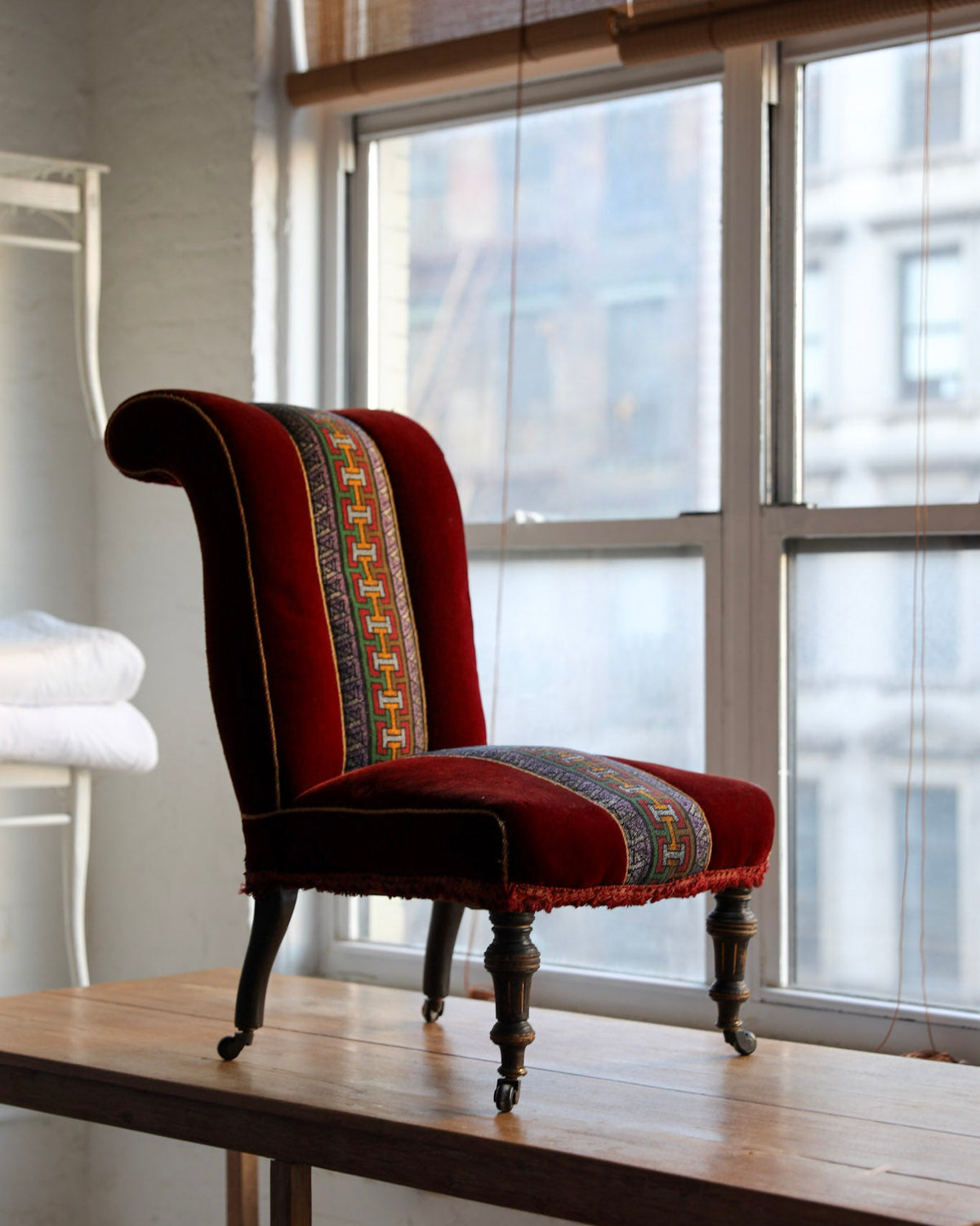 A vintage-style wingback armchair upholstered in deep red velvet with a colorful patterned central panel, shown on a wooden table in front of a bright window.