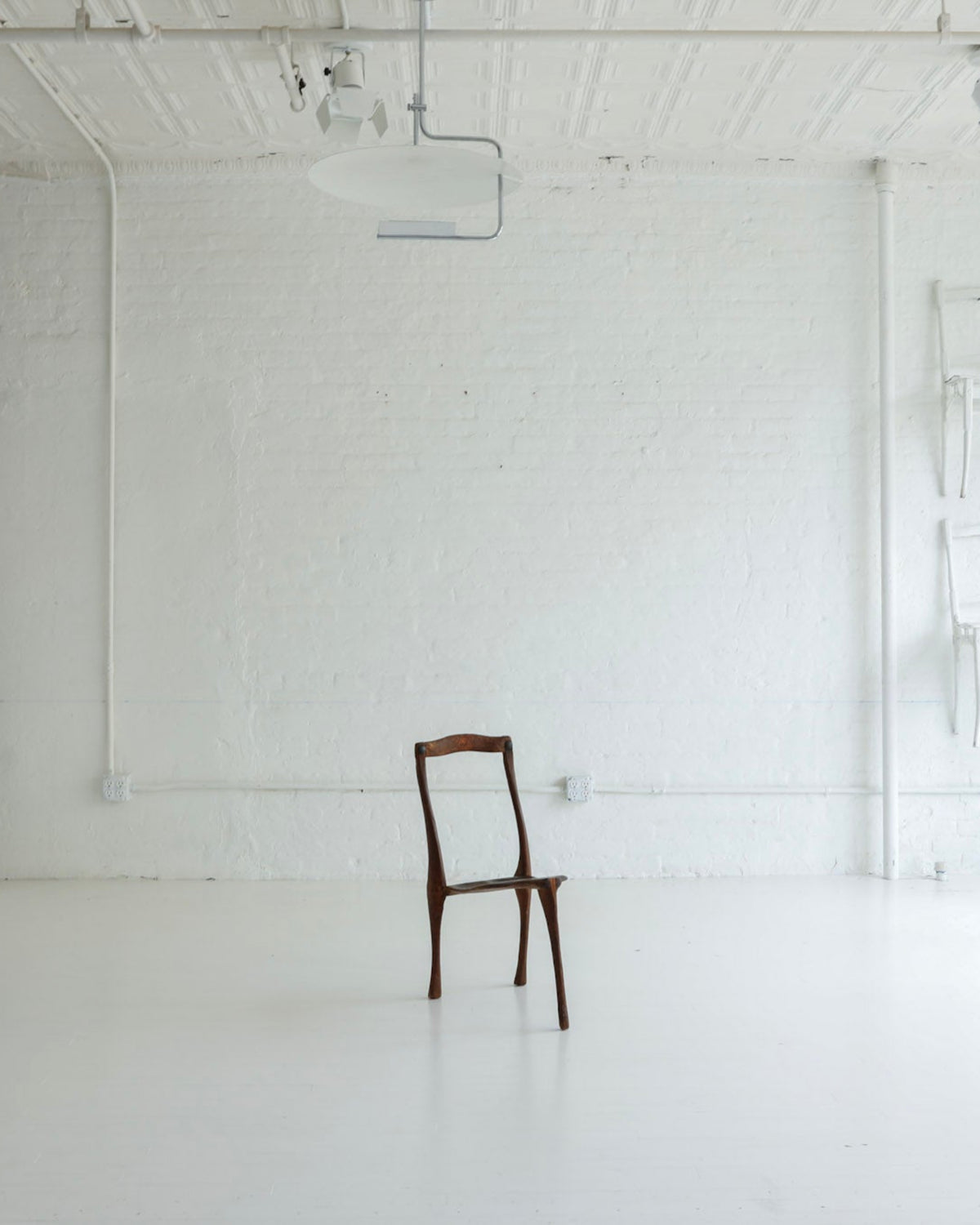 A minimalist white studio or gallery space with a single dark wooden chair centered on a white floor, alongside white shelving and wall-mounted chairs near a large window.