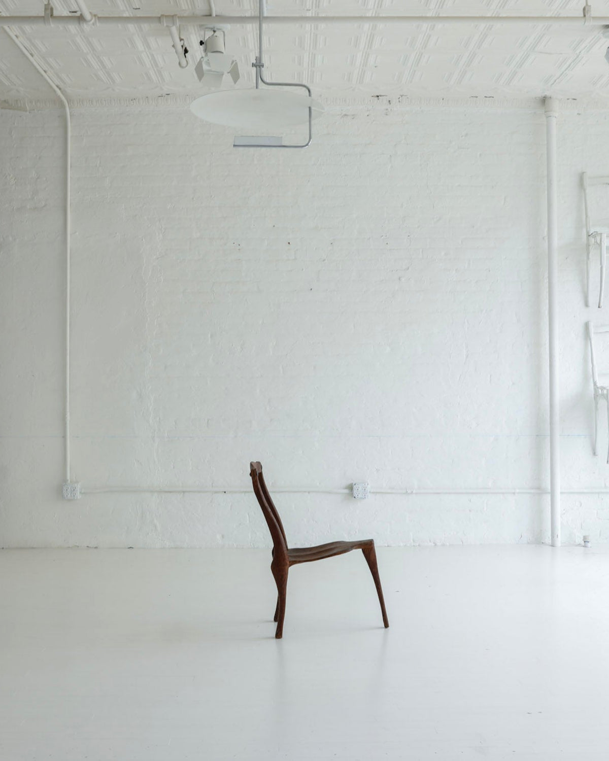 A single dark brown wooden chair sits centered in a bright, minimalist white studio or gallery space with white brick walls, exposed ceiling pipes, and several white chairs mounted on the wall to the right.