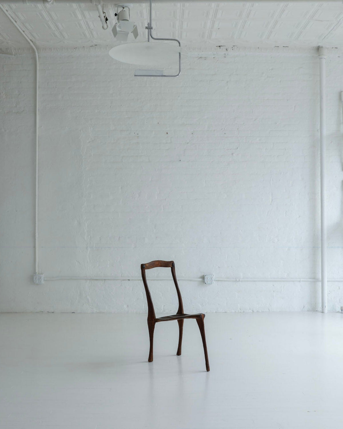 A minimalist art gallery or studio with white brick walls and a white floor, featuring a single dark brown wooden chair centered in the room, with other white chairs mounted on the walls and a white cabinet on the left.