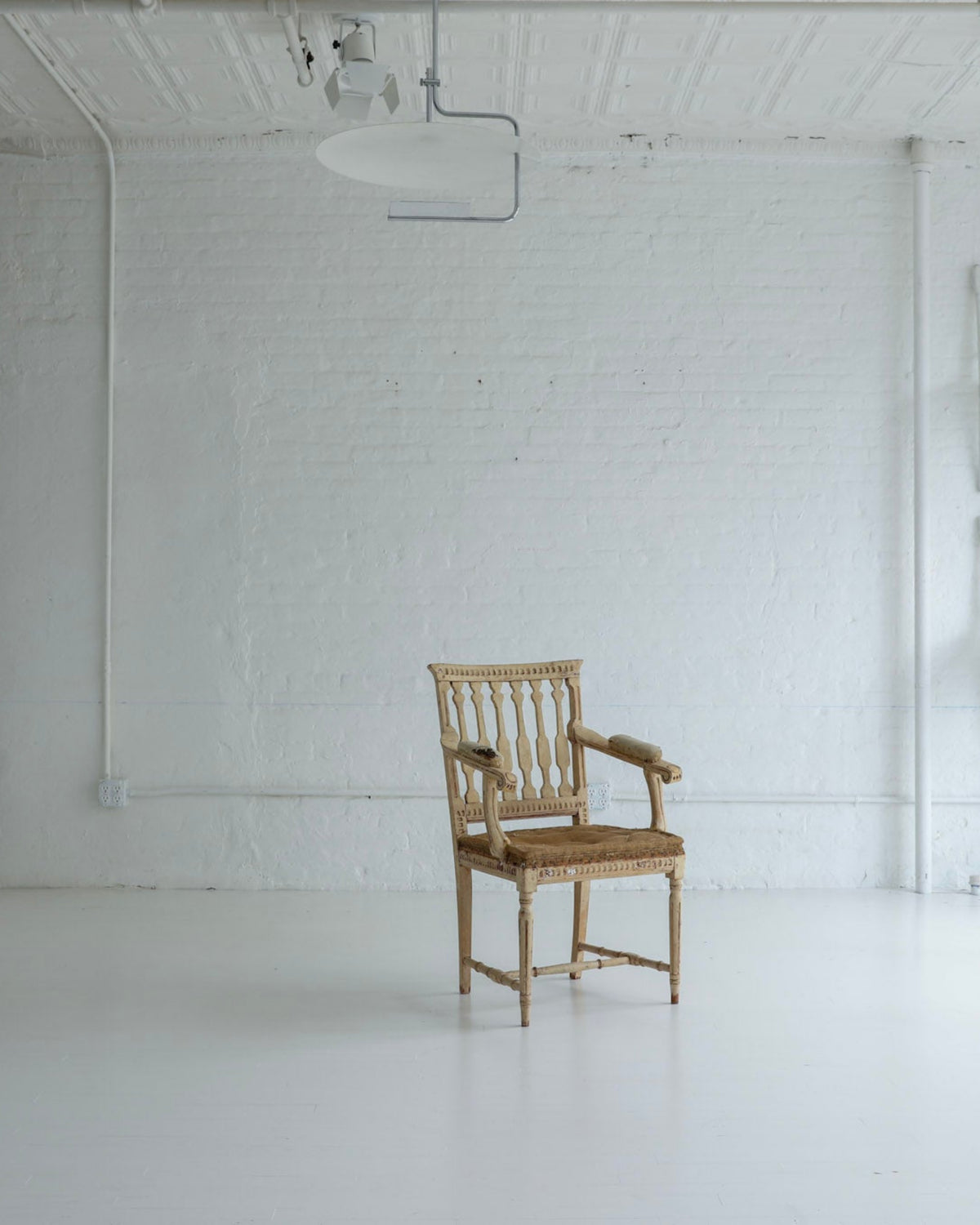 A single light brown wooden armchair sits centered in a bright, white studio or gallery space with white brick walls, track lighting, and additional white chairs mounted on the wall.