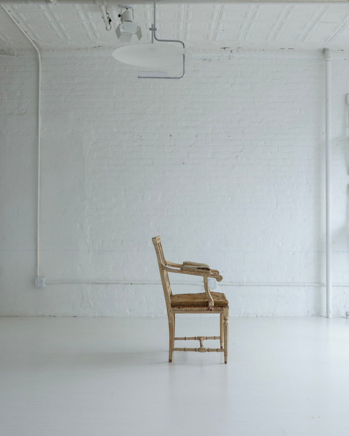 A rustic wooden armchair sits in the center of a bright, minimalist white studio space with white brick walls, industrial ceiling tracks, and other white chairs arranged against the background.