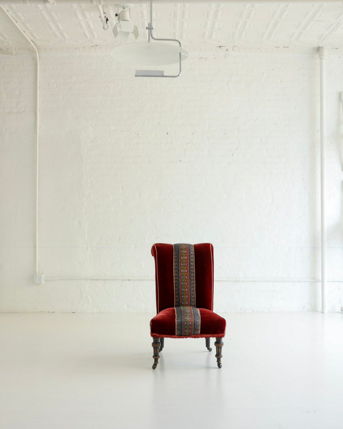 A single red upholstered armchair with a striped patterned backrest stands centered in a bright white studio or gallery space with white walls, ceiling lights, and additional white furniture pieces in the background.