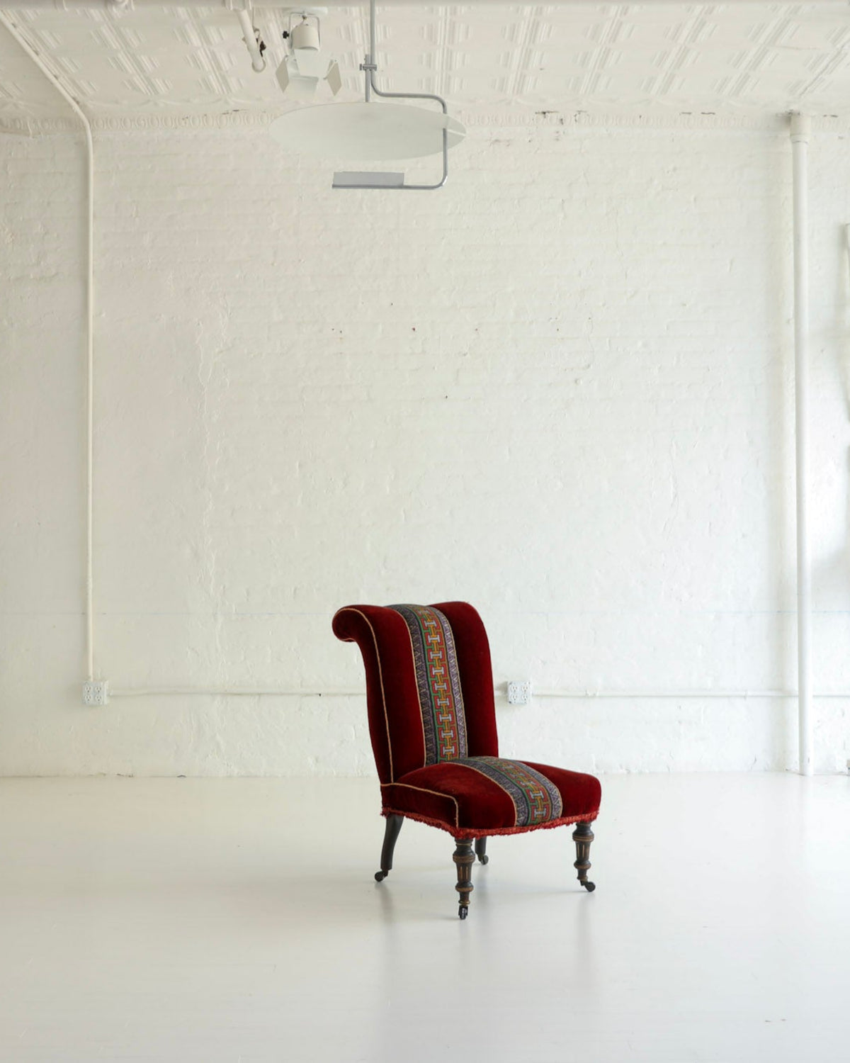 A single upholstered armchair in deep red velvet with a patterned striped backrest and dark wooden legs, placed in a bright white studio or gallery space with white walls, ceiling lights, and other white furniture pieces in the background.