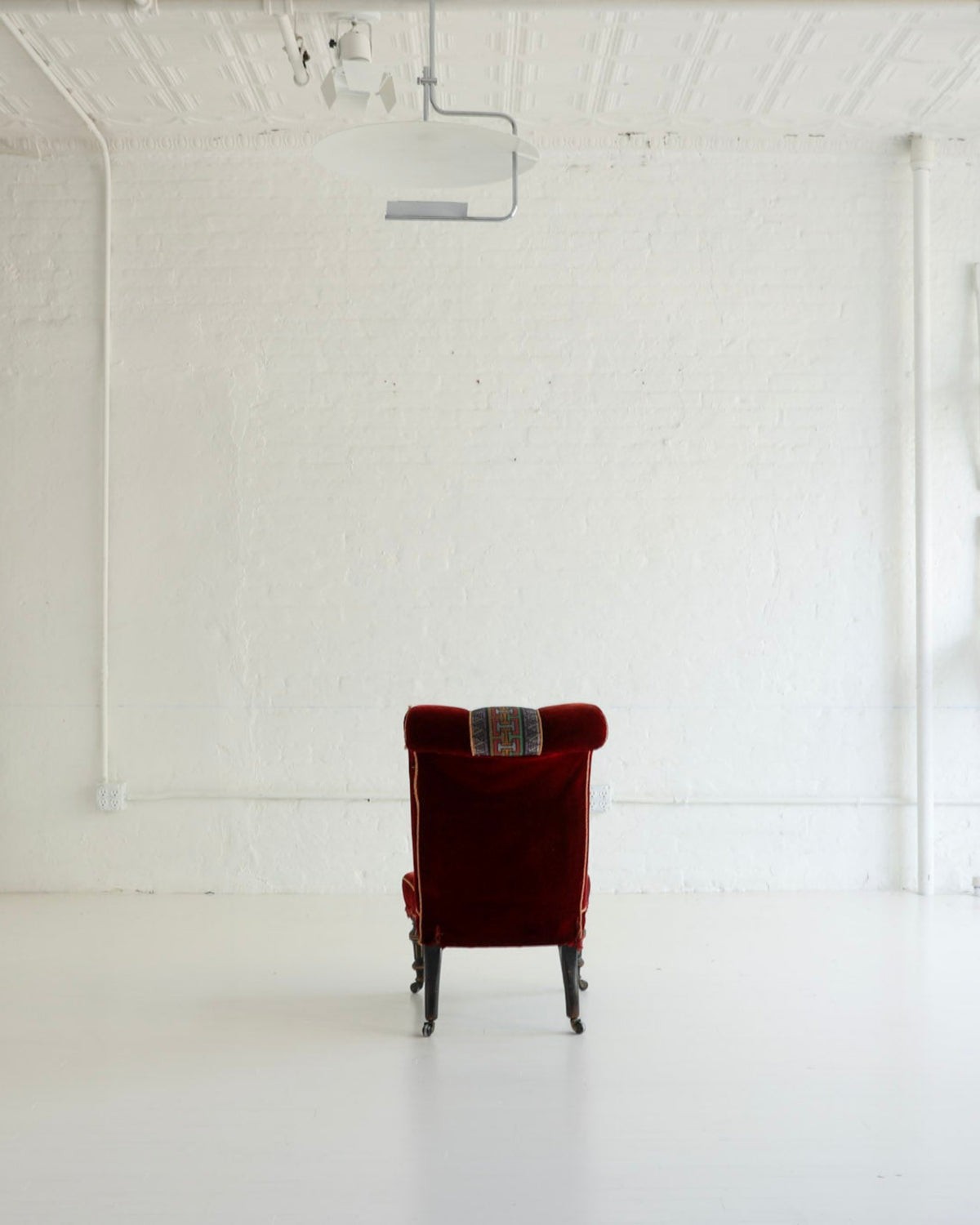 A single deep red velvet armchair is centered in a bright, white studio gallery space with brick walls, track lighting, and white chairs mounted on the wall.