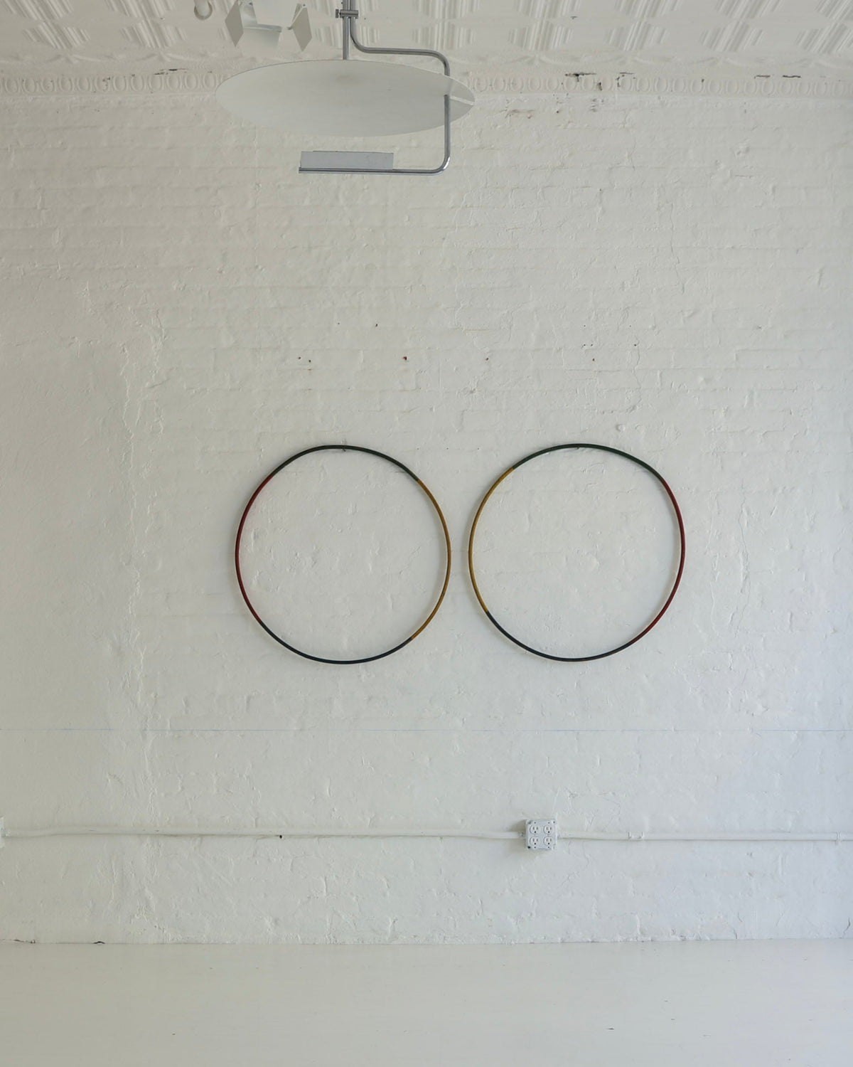 Two circular wall art pieces with thin black, red, and gold-toned metal rings mounted on a white painted brick wall in a gallery or studio space.