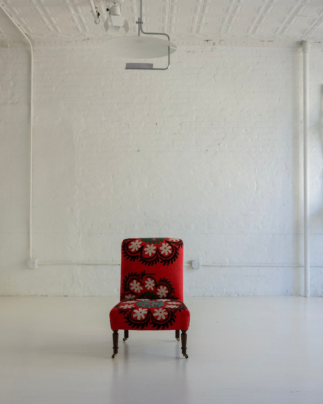 A single red upholstered armchair with a floral pattern sits centered in a bright white studio or gallery space with white walls, ceiling, and floor. Several white chairs are stacked against the wall on the right.