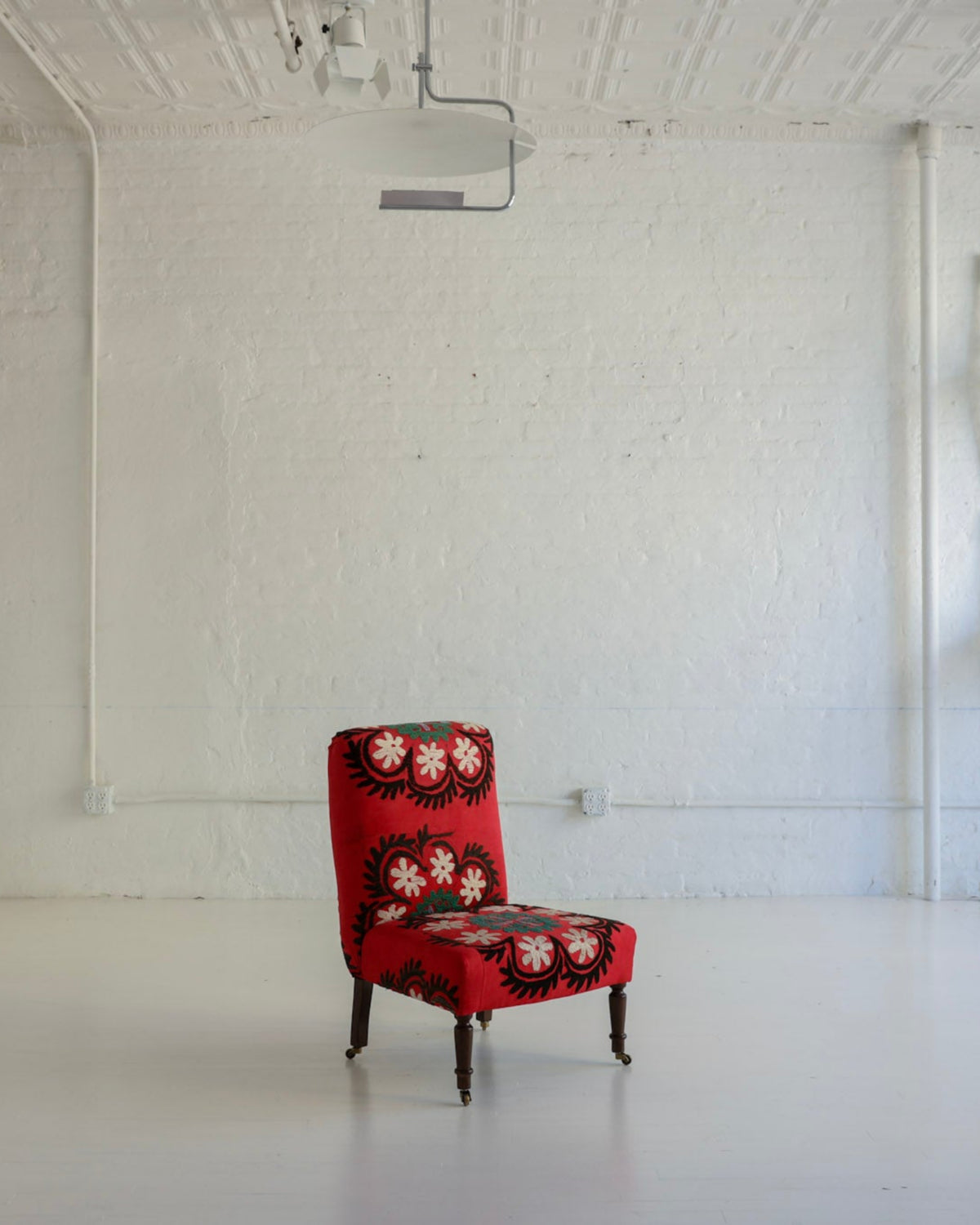 A single upholstered armchair with a red base and black-and-white floral pattern sits in the center of a bright, white studio or gallery space with exposed ceiling beams and wall-mounted white chairs.
