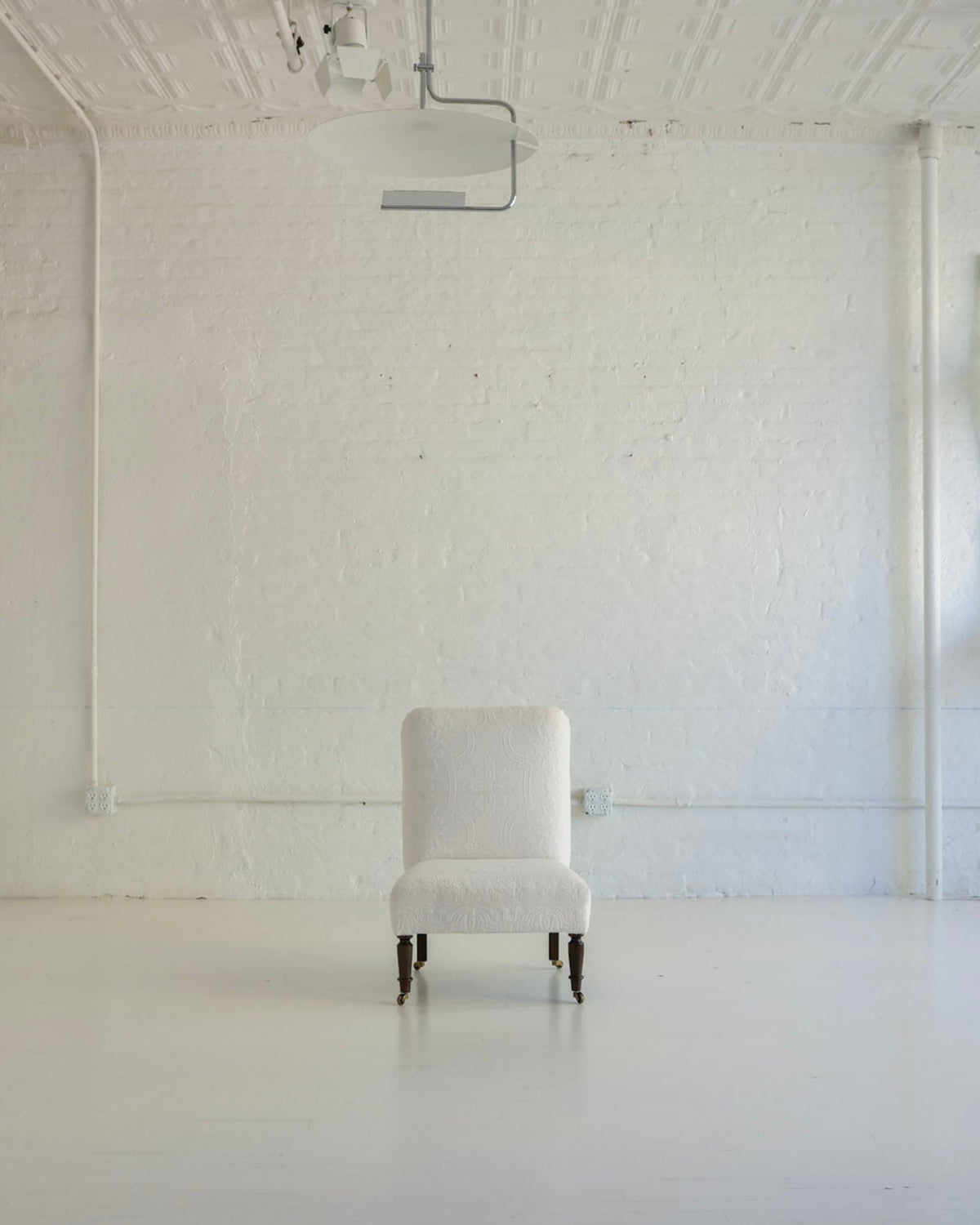 A minimalist white upholstered armchair sits centered in a bright, white-painted studio room with exposed brick walls, track lighting, and additional white chairs stacked against the right wall.