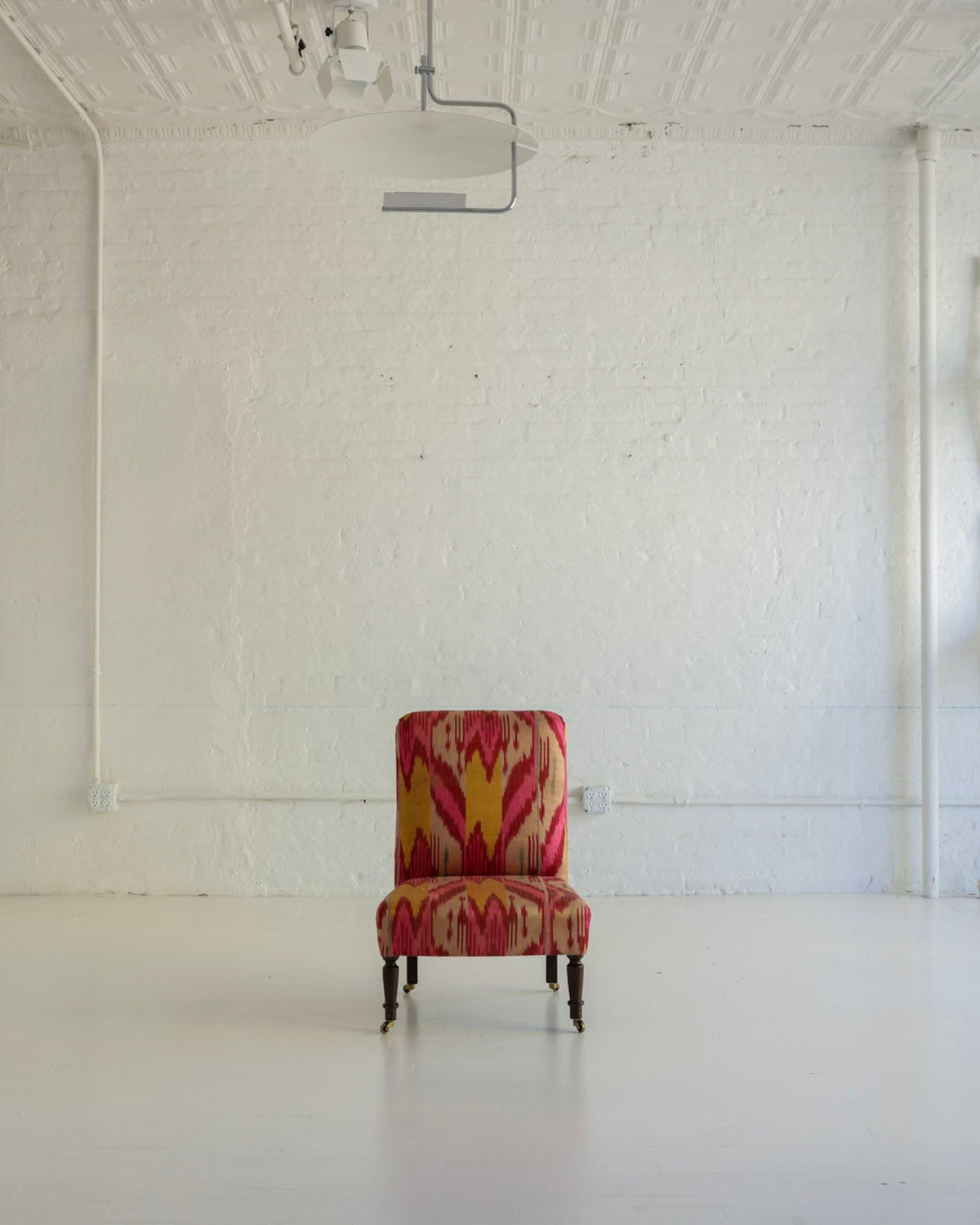 A single upholstered accent chair with a red, orange, and yellow geometric pattern sits centered in a bright white studio or gallery space with white walls, a projector on the ceiling, and several white chairs mounted on the wall.