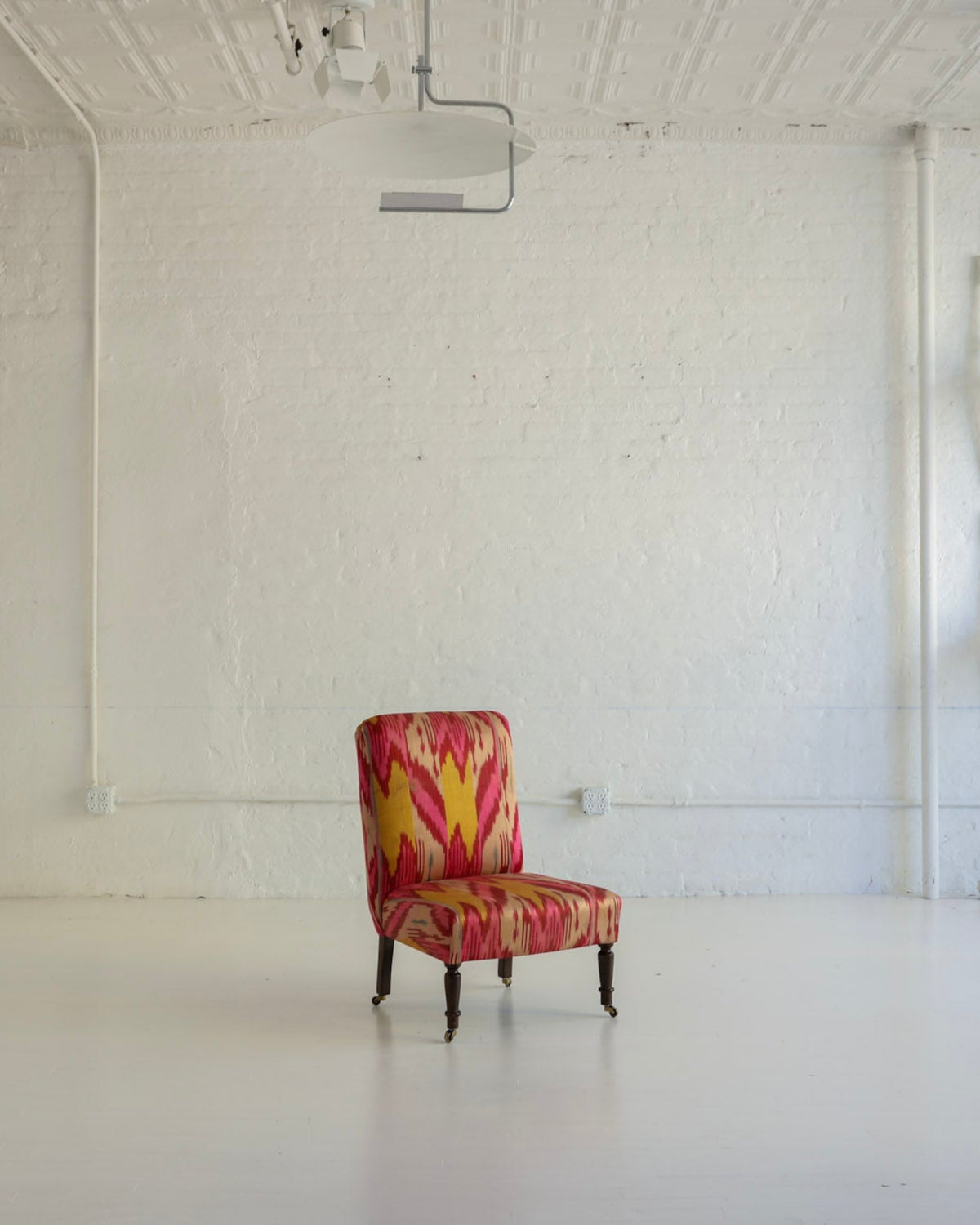 A single upholstered armchair with a bold red, orange, and yellow floral pattern sits in the center of a bright, white studio or gallery space with white walls, a projector on the ceiling, and several white chairs mounted on the wall.