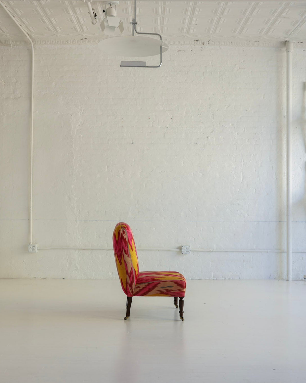 A single upholstered accent chair with a red, orange, and yellow abstract pattern sits centered in a bright, white studio or gallery space with exposed ceiling beams and several white chairs mounted on the wall.