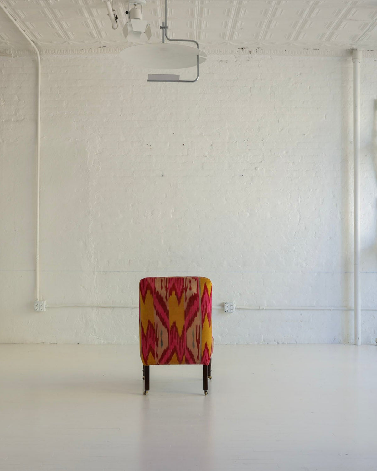 A single upholstered armchair with a bold red, orange, and yellow geometric pattern sits centered in a bright, white studio or gallery space with exposed brick walls, white chairs mounted on the wall, and a projector hanging from the ceiling.