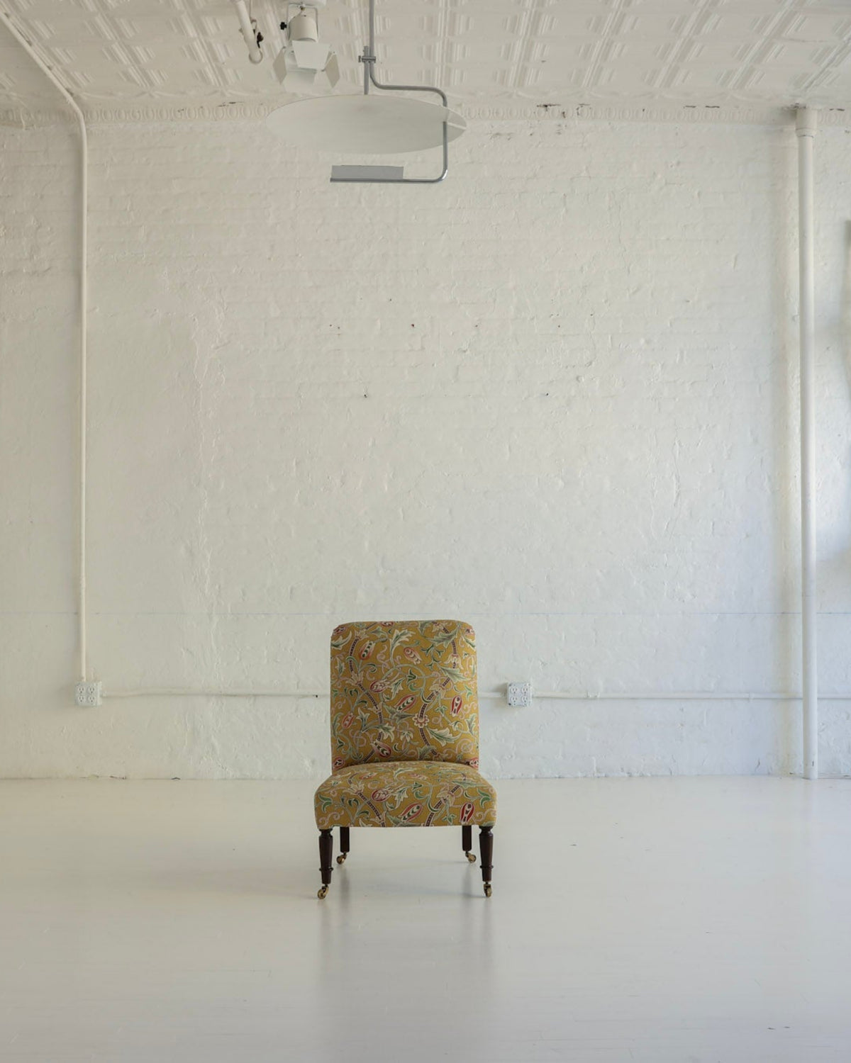 A single upholstered armchair with a dense floral pattern in muted yellow, green, and orange sits centered in a bright, white studio or gallery space. The room has white brick walls, a white ceiling with track lighting, and several white chairs stacked against the right wall.