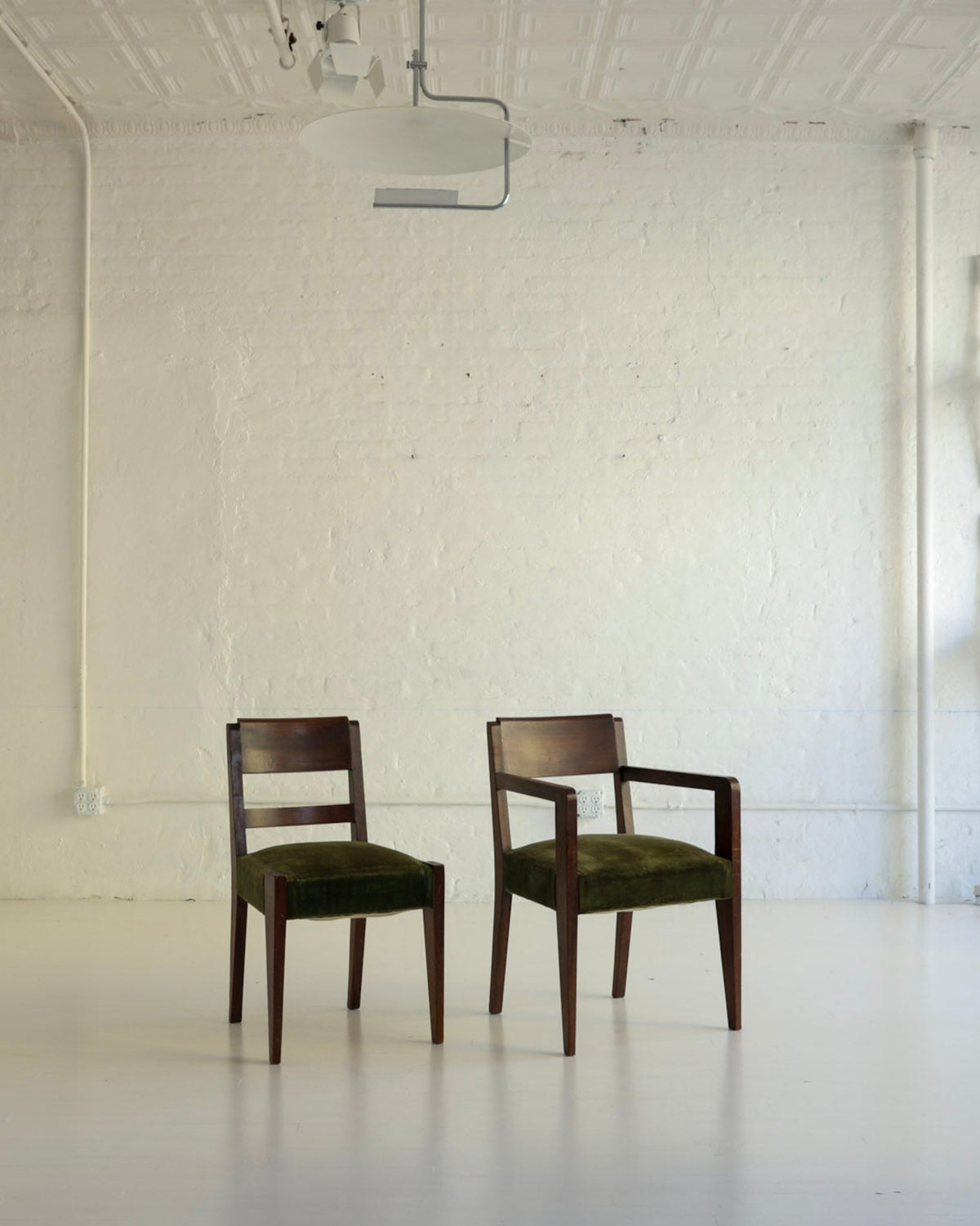 Two wooden armchairs with dark green upholstered seats are placed in a bright, white studio or gallery space with exposed brick walls, white pipes, and a projector on the ceiling. A set of six white chairs is stacked against the right wall.