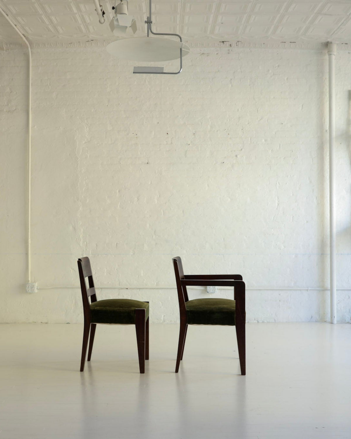 A minimalist interior room with white walls and floor, featuring two dark wooden chairs with olive green seats positioned centrally. Additional white chairs are stacked against the wall on the right, and a tall white cabinet or partition stands on the left.