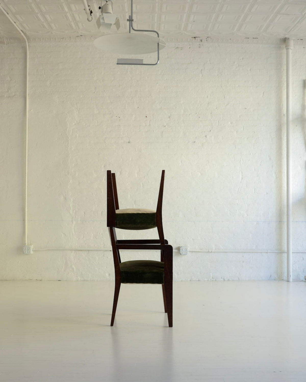A dark brown wooden chair with green upholstered seat and back cushions stands centered in a bright, white studio or gallery space with white brick walls, track lighting, and additional chairs mounted on the wall.