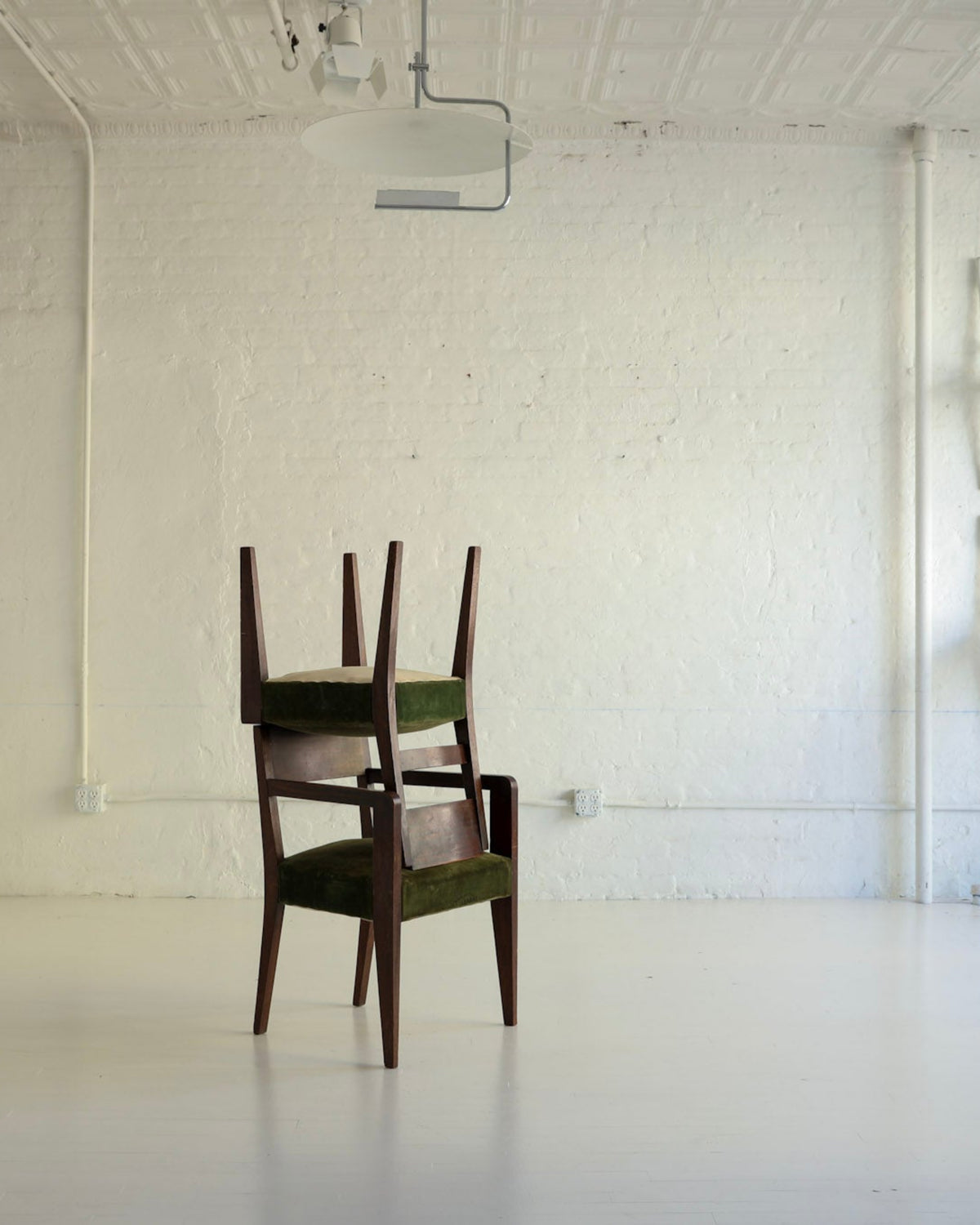 A dark wood chair with green upholstered seat and back is centered in a bright white studio or gallery space with exposed brick walls, track lighting, and additional chairs mounted on the wall.