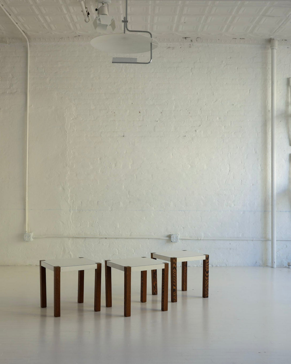 A minimalist white studio or gallery space with a white brick wall, white floor, and several small wooden stools arranged in a row near the center. A set of four white chairs is stacked and hung on the right wall.
