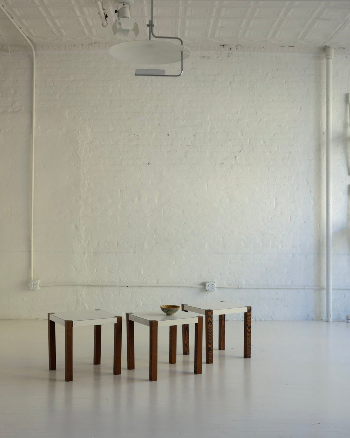 A minimalist white studio or gallery space with a white brick wall, white floor, and several white chairs arranged against the wall, alongside a few small wooden-and-white stools in the center.