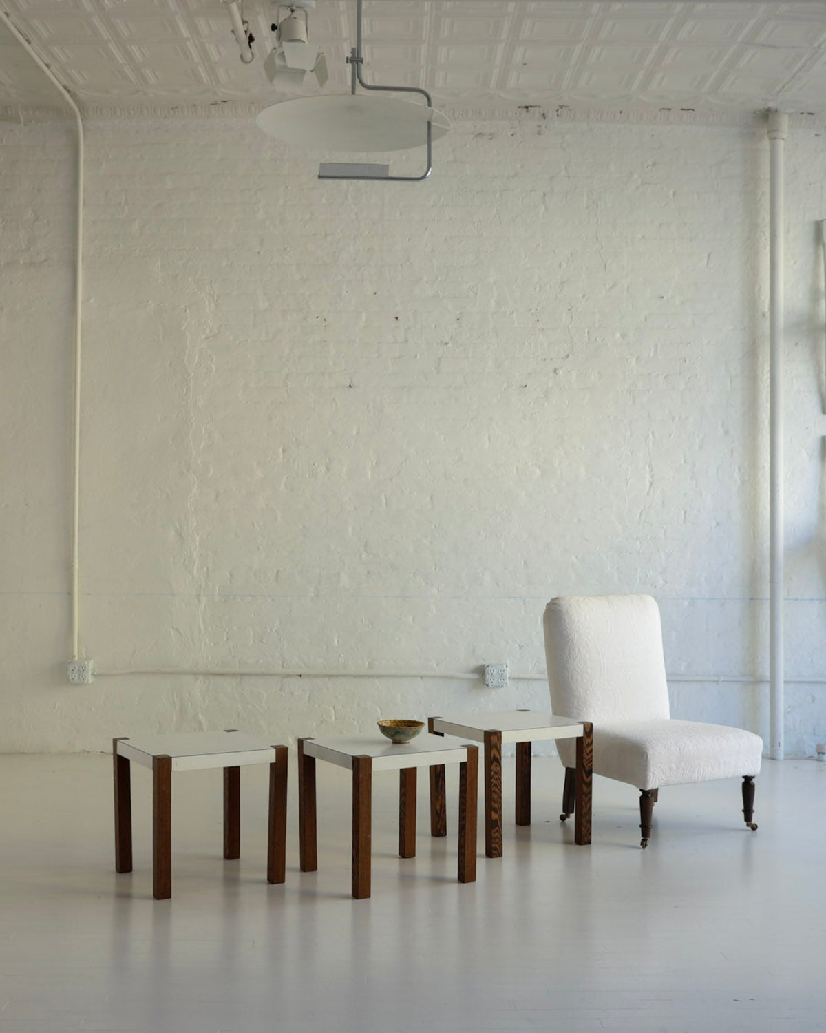 A minimalist white brick room with a white upholstered armchair and four small matching side tables, alongside a tall white cabinet and a stack of chairs on the wall.