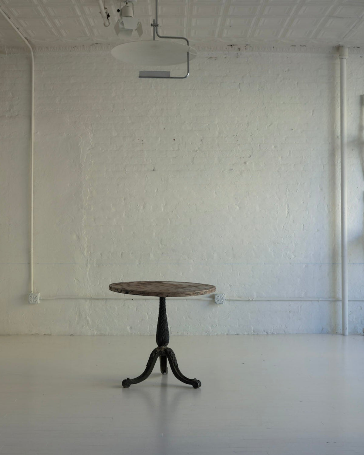 A minimalist interior room with white walls and floor, featuring a small round wooden table centered in the foreground. Several white chairs are stacked or hung against the right wall, and a tall white cabinet or screen stands on the left.