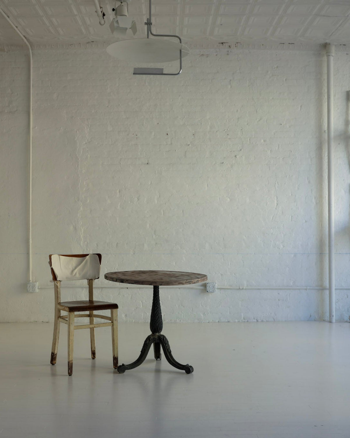 An empty white studio room with a small round table and chair in the center, additional chairs stacked against the wall, and a tall white cabinet on the left.
