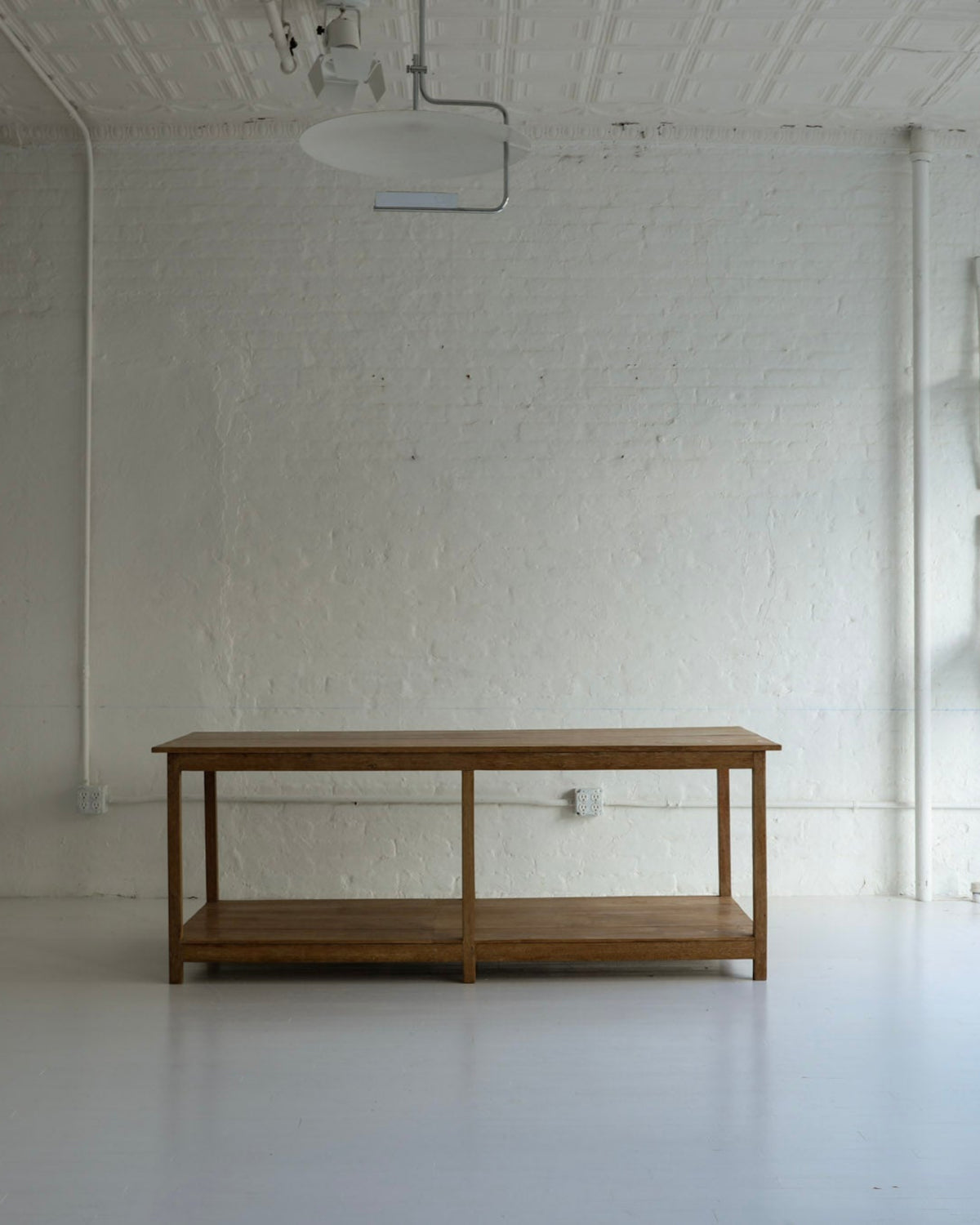 A minimalist wooden table with two shelves stands in a bright, white studio or gallery space with white brick walls, track lighting, and stacked white chairs on the wall.