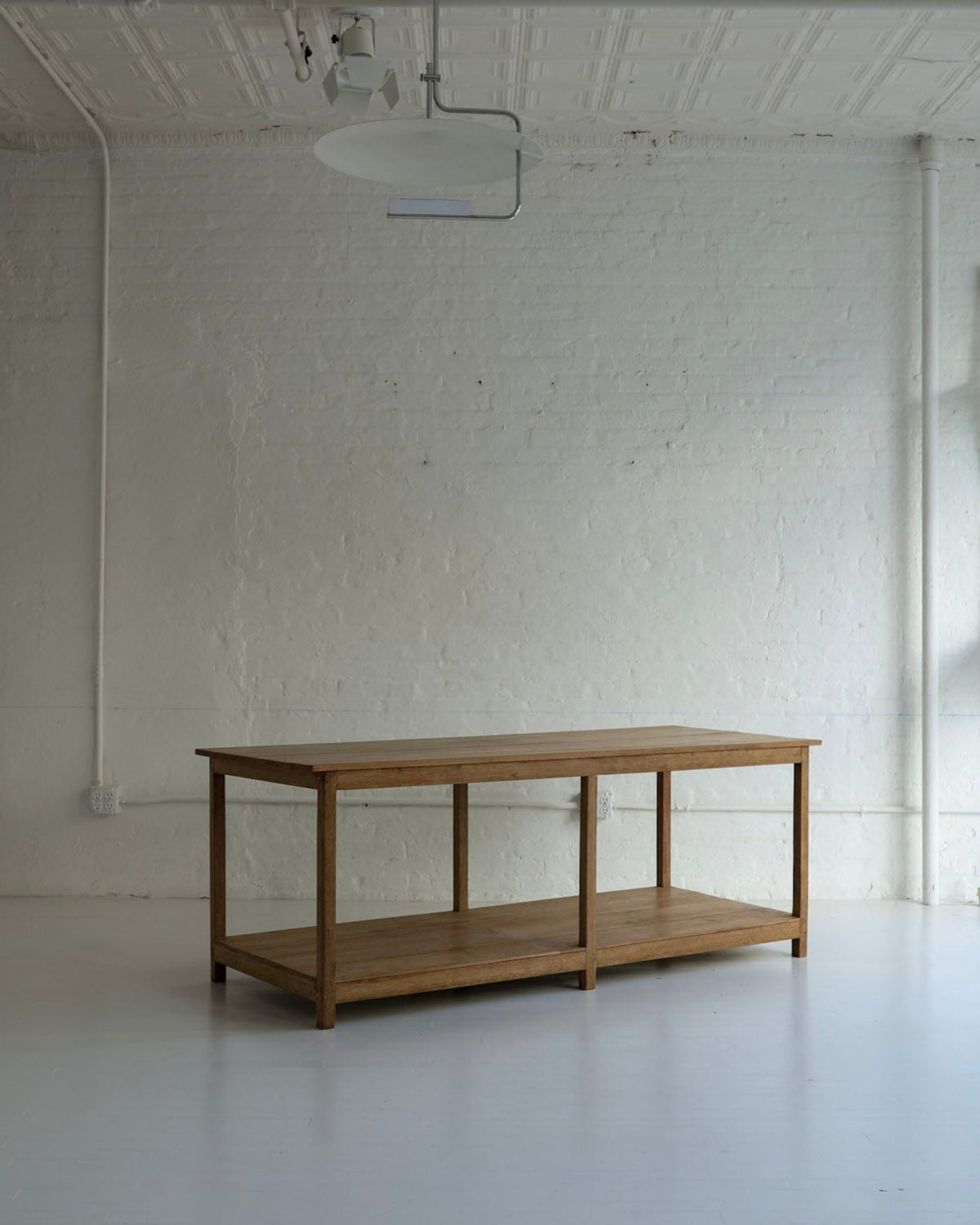 A wooden two-tier table or console stand with a natural oak finish, positioned in a bright, minimalist white studio room with brick walls.