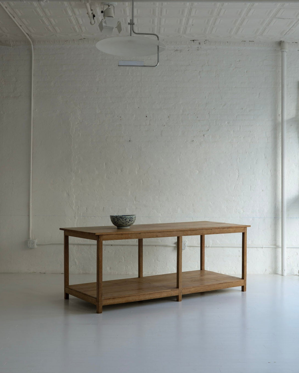 A wooden two-tier table with a bowl on top, set in a bright, white studio or gallery space with white brick walls, hanging chairs, and a projector on the ceiling.