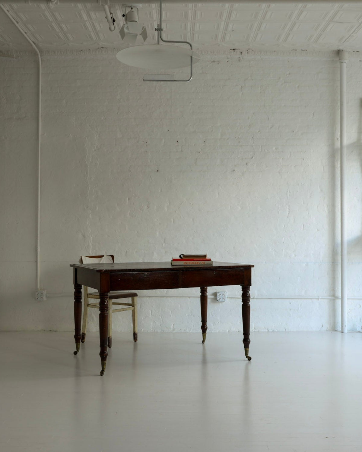 A large, minimalist white room with a dark wooden writing desk in the center, a matching chair, a tall white cabinet on the left, and four white chairs stacked against the right wall.