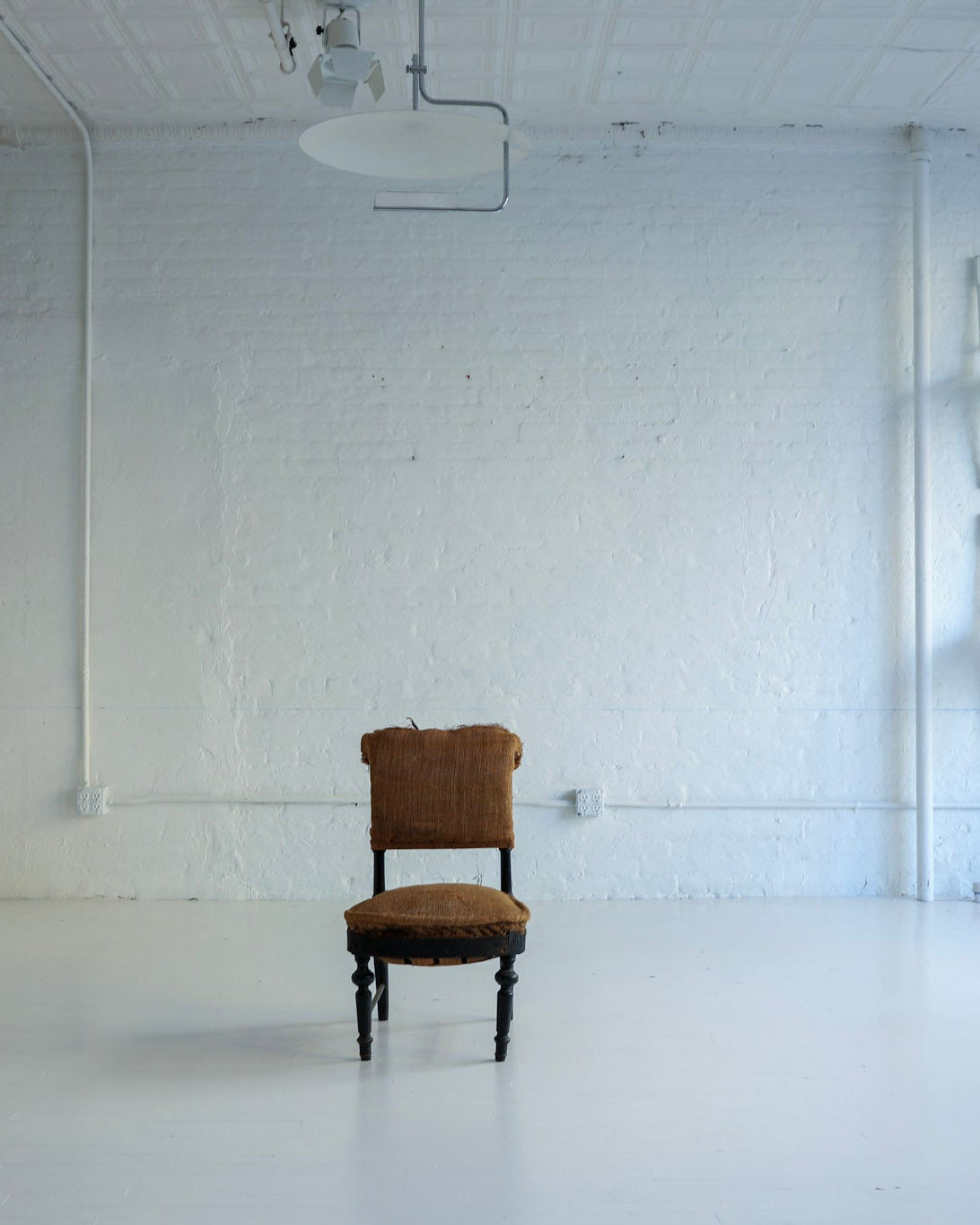 A single brown upholstered wooden chair stands centered in a bright white studio or gallery space with white brick walls, white floor, hanging lights, and several white chairs stacked against the right wall.