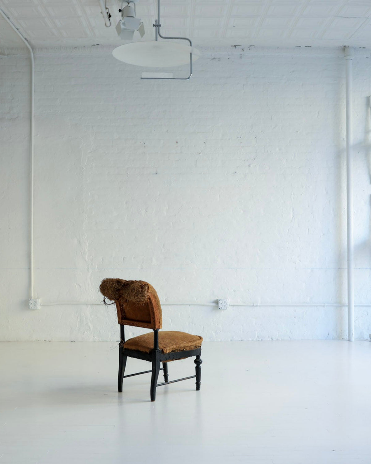 A single brown upholstered chair with a dark wooden frame sits in the center of a bright, white studio or gallery space with white brick walls, white floor, and several white chairs mounted on the wall.