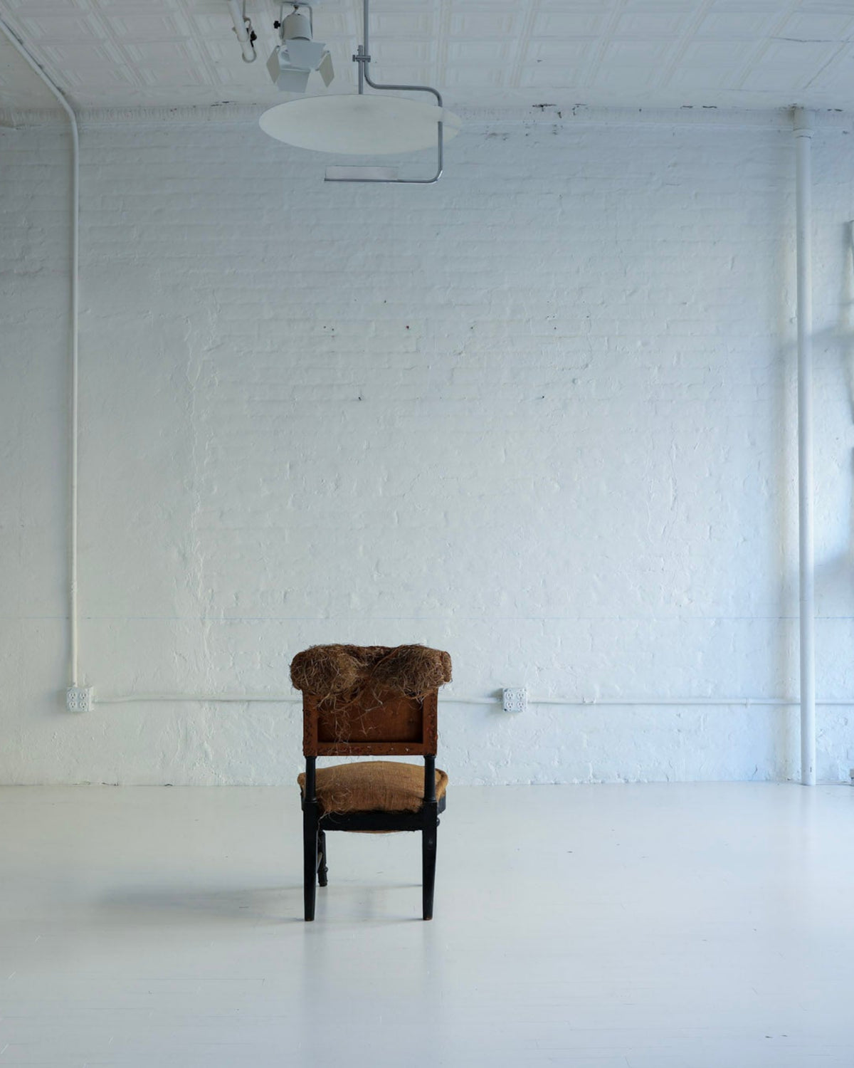 A vintage brown armchair with a brown fur-like collar is centered in a bright, white-painted studio room with industrial ceiling fixtures and white chairs mounted on the wall.